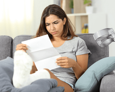 Woman with cast on her leg propped up on coffee table looks puzzled at a piece of paper