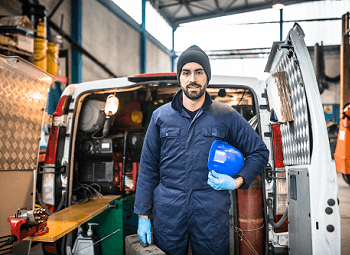 Man wearing work jumpsuit and holding a hard hat stands in front of the open back of a work van